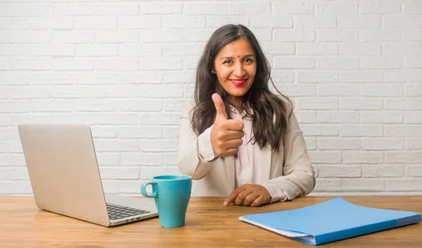Young indian woman at the office cheerful and excited, smiling and raising her thumb up, concept of success and approval, ok gesture