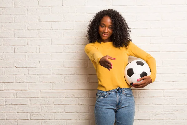 Young black woman with soccer ball reaching out to greet someone against brick wall