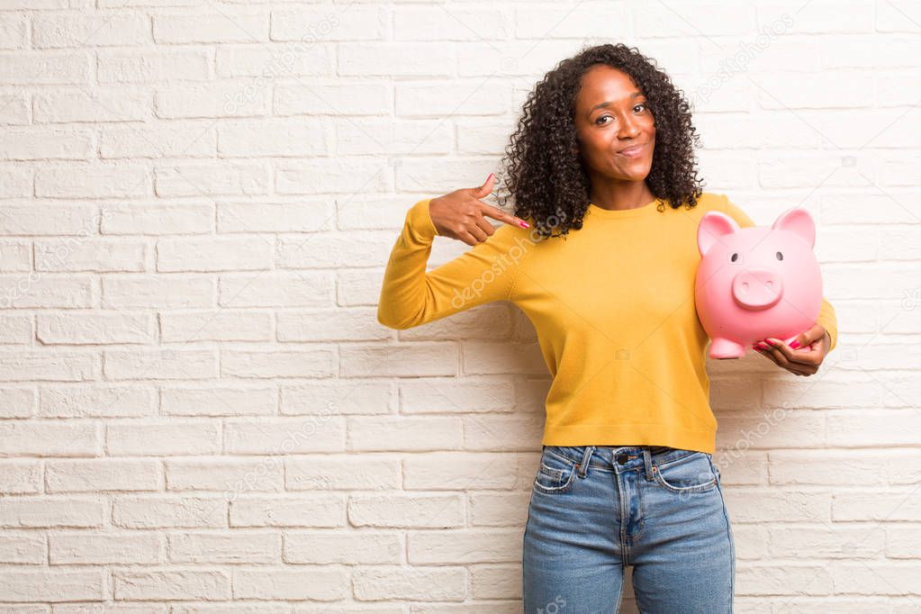 Young black woman with piggy bank proud and confident, pointing finger against brick wall