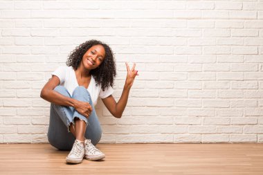 Ahşap zemin eğlenceli ve mutlu, pozitif ve doğal, üzerinde oturan genç siyah kadın yapar duvara karşı zafer bir jesti