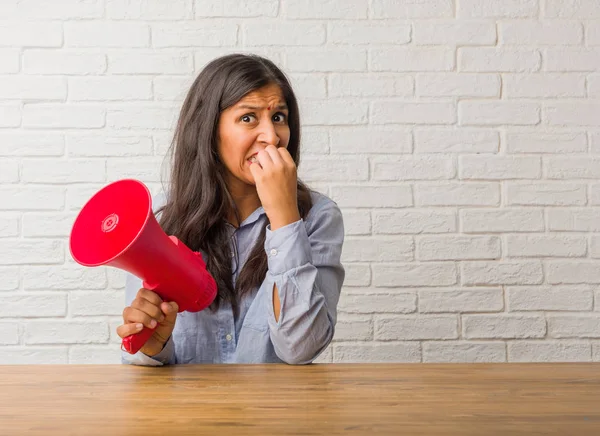 Young indian woman biting nails, nervous and very anxious and scared for the future, feels panic and stress. Holding a megaphone.