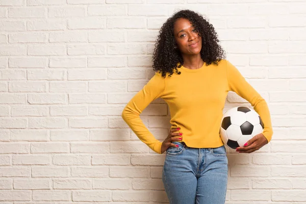レンガの壁に腰に手をサッカー ボールで若い黒人女性 — ストック写真
