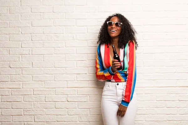 Young black woman in colorful shirt with beer bottle looking up against brick wall