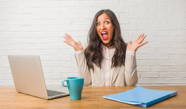 Young indian woman at the office screaming happy, surprised by an offer or a promotion, gaping, jumping and proud