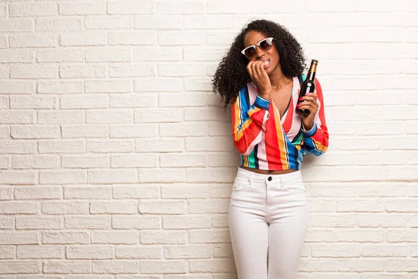 Young black woman in colorful shirt with beer bottle biting nails against brick wall
