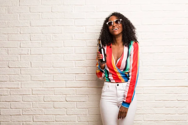 Young black woman in colorful shirt with beer bottle looking up against brick wall