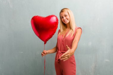 Birisi ya da el hareketi, mutlu ve heyecanlı yardımcı olmak için selamlıyorum uzanarak zarif sarışın genç kadın portresi. Bir kırmızı kalp balon holding.