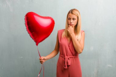 Portre sarışın genç zarif kadın şüphe ve karışık, düşünme, bir fikir ya da bir şey hakkında endişeli. Bir kırmızı kalp balon holding.