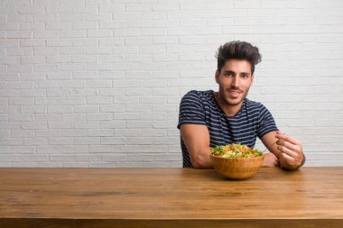 Kalça, rahat ve gülümseyen, çok olumlu ve neşeli ayakta, el ile bir masada oturan genç yakışıklı ve doğal adam. Taze bir salata yemek.