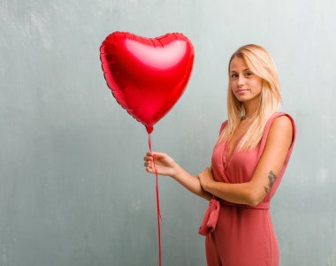 Kollarını, gülümseyen ve mutlu crossing, kendinden emin ve dostu zarif sarışın genç kadın portresi. Bir kırmızı kalp balon holding.