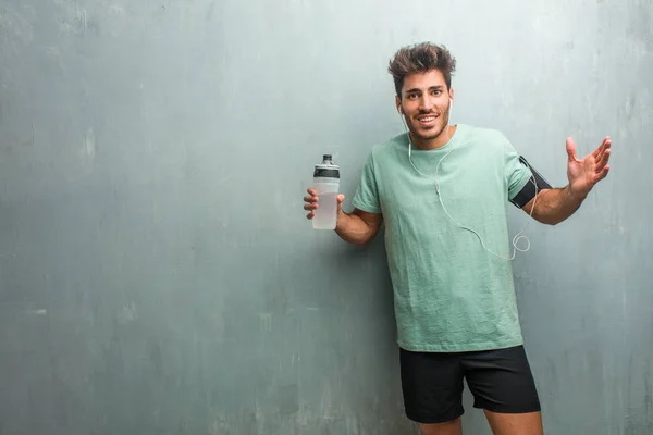 Young fitness man with sports bottle against a grunge wall screaming happy, wearing an armband with phone.