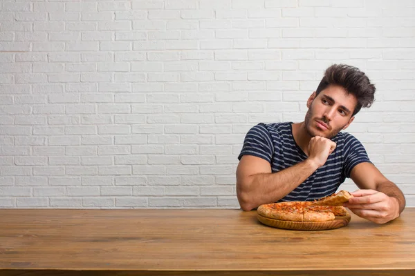 Young handsome and natural man sitting on a table thinking and looking up, confused about an idea, would be trying to find a solution. Eating a delicious pizza.