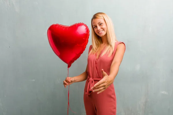 Portrait of young elegant blonde woman reaching out to greet someone or gesturing to help, happy and excited. Holding a red heart balloon.