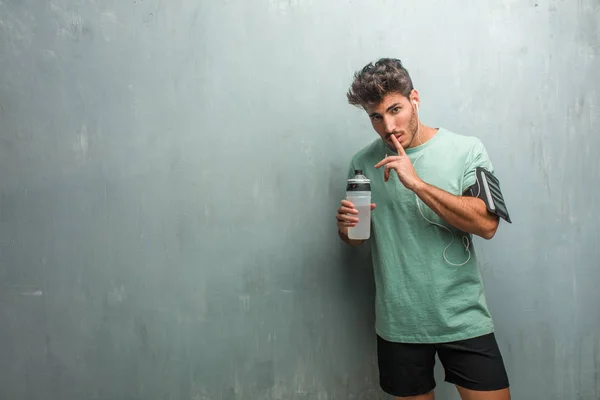Young fitness man against a grunge wall with sports bottle keeping a secret or asking for silence, wearing an armband with phone.