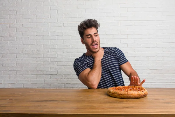 Young handsome and natural man sitting on a table worried and overwhelmed, anxious feeling pressure, concept of anguish. Eating a delicious pizza.