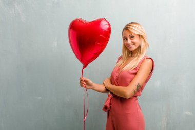 Kollarını, gülümseyen ve mutlu crossing, kendinden emin ve dostu zarif sarışın genç kadın portresi. Bir kırmızı kalp balon holding.