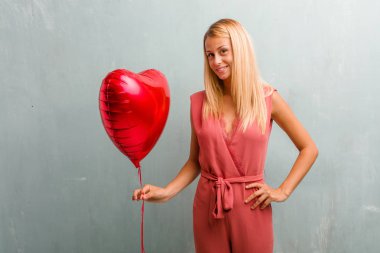 Ellerle kalça, ayakta, rahat ve gülümseyen, çok olumlu ve neşeli sarışın genç zarif kadın portresi. Bir kırmızı kalp balon holding.