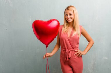 Ellerle kalça, ayakta, rahat ve gülümseyen, çok olumlu ve neşeli sarışın genç zarif kadın portresi. Bir kırmızı kalp balon holding.