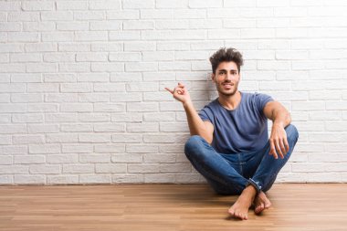 Tarafına işaret eden bir ahşap zemin üzerinde oturup sürpriz bir şey, doğal ve rahat sunan gülümseyen genç doğal adam