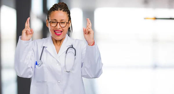 Portrait of a young black doctor woman crossing his fingers, wishes to be lucky for future projects, excited but worried, nervous expression closing eyes