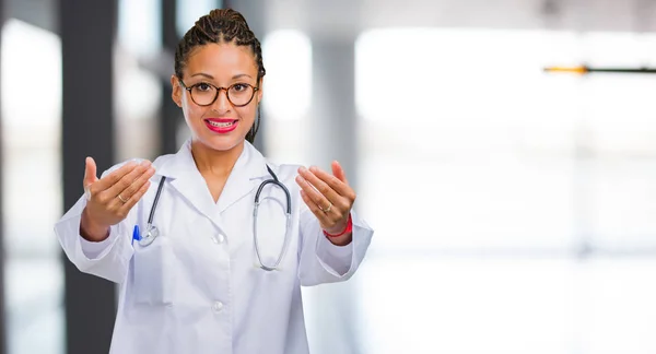 Portrait of a young black doctor woman inviting to come, confident and smiling making a gesture with hand, being positive and friendly