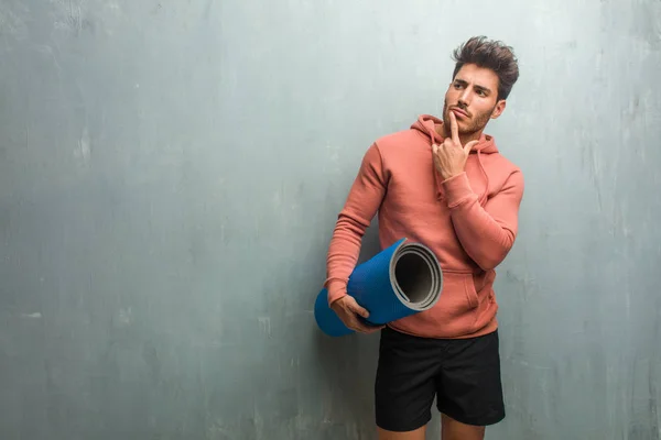 Young fitness man against a grunge wall doubting and confused, thinking of an idea or worried about something. Holding a blue mat for practicing yoga.