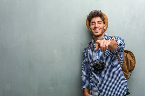 Young handsome traveler man wearing a straw hat, a backpack and a photo camera shouting, laughing and making fun of another, concept of mockery and uncontrol