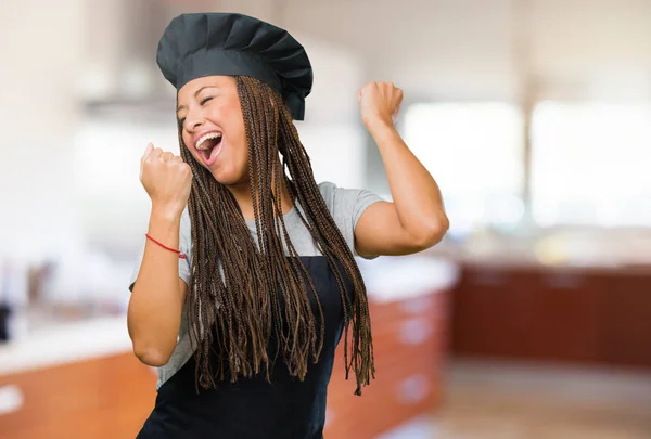 Portrait of a young black baker woman very happy and excited, raising arms, celebrating a victory or success, winning the lottery