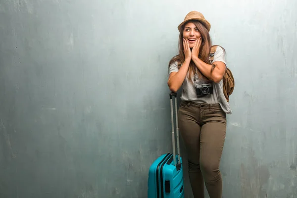 Portrait of young traveler latin woman against a wall surprised and shocked, looking with wide eyes, excited by an offer or by a new job, win concept. Holding a blue suitcase.