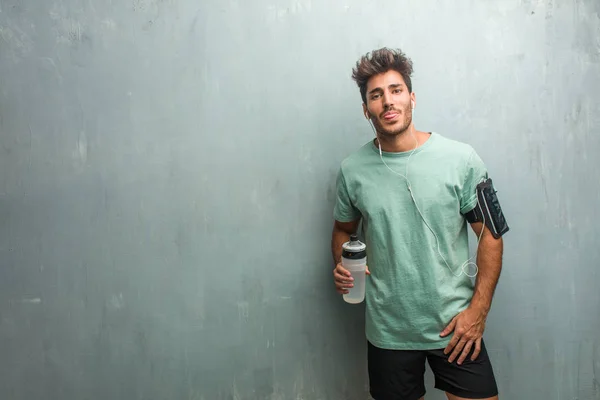 Young fitness man with sports bottle against a grunge wall expression of confidence and emotion, wearing an armband with phone.