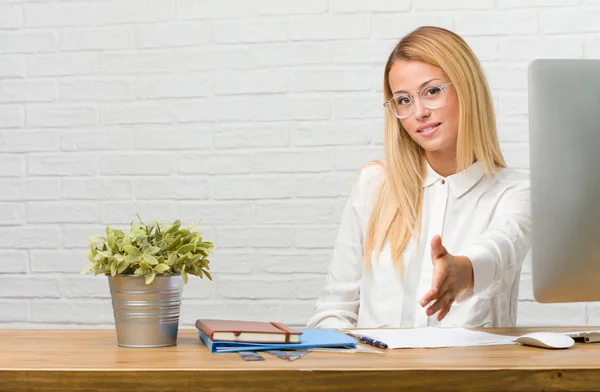 Portrait of young student sitting on her desk doing tasks reaching out to greet someone or gesturing to help, happy and excited