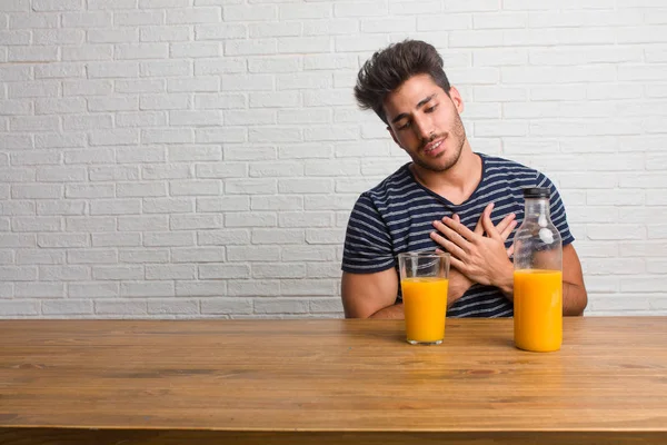 年轻的英俊和自然的男人坐在桌子上做一个浪漫的手势 爱上一个人或显示对一些朋友的爱 包括橙汁和麦片碗 图库图片