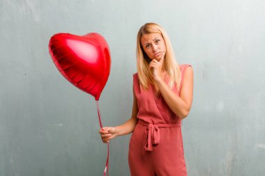 Portre sarışın genç zarif kadın şüphe ve karışık, düşünme, bir fikir ya da bir şey hakkında endişeli. Bir kırmızı kalp balon holding.