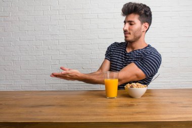 Elleriyle bir şey tutan, bir ürün, gülümseyen ve neşeli gösterilen, hayali bir nesne sunan bir masada oturan genç yakışıklı ve doğal adam. Bir kahvaltı yapıyor, portakal suyu ve tahıl bowl içerir.