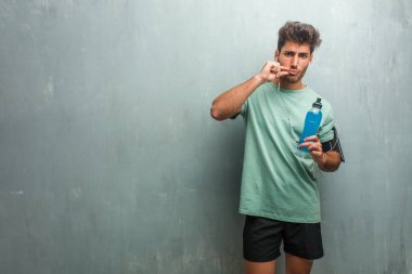 Bir mavi enerji içeceği tutan genç fitness adam gizli tutmak veya sessizlik için soran bir grunge duvara.