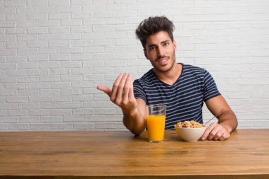 Pozitif ve güler yüzlü gel, kendinden emin ve gülümseyen bir hareketi el ile yapmak için davet bir masada oturan genç yakışıklı ve doğal bir adam. Bir kahvaltı yapıyor, portakal suyu ve tahıl bowl içerir.