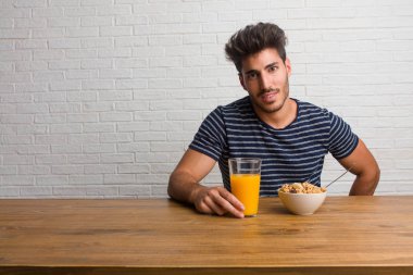 Kalça, rahat ve gülümseyen, çok olumlu ve neşeli ayakta, el ile bir masada oturan genç yakışıklı ve doğal adam. Bir kahvaltı yapıyor, portakal suyu ve tahıl bowl içerir.