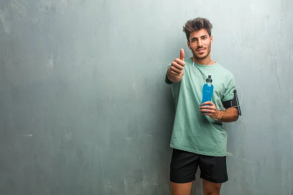 Young fitness man against a grunge wall cheerful and excited, ok gesture, holding a blue energy drink.