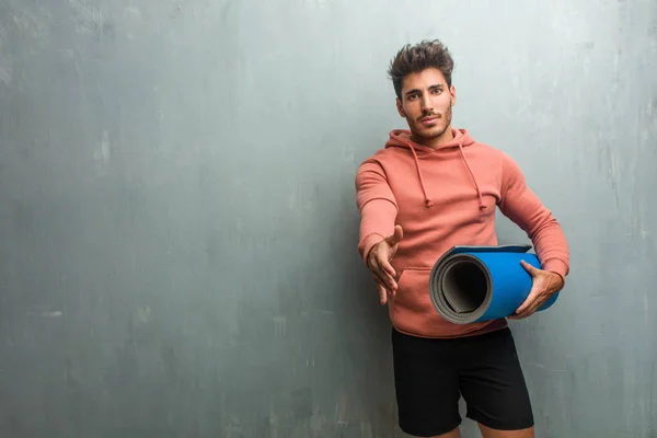 Young fitness man against a grunge wall reaching out to greet someone or gesturing to help, happy and excited. Holding a blue mat for practicing yoga.
