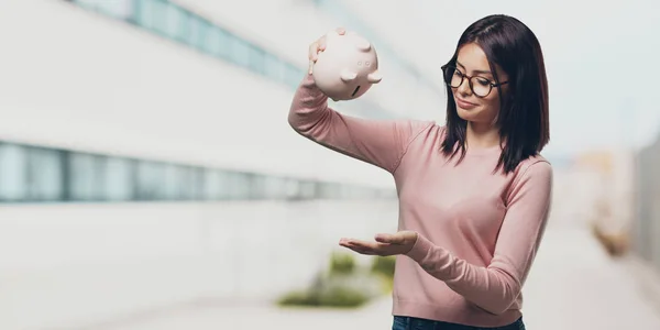 Young pretty woman sad and disappointed, holding a piglet bank, no money left, trying to get something out, face of anger and anguish, concept of poverty in the university campus