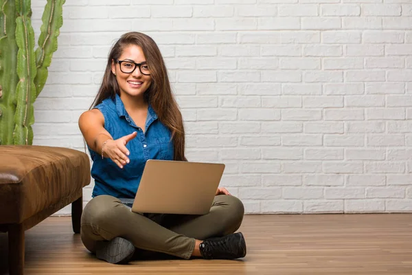 Portrait of young latin woman sitting on the floor reaching out to greet someone or gesturing to help, happy and excited. Holding a laptop.