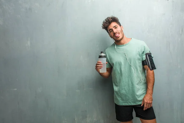 Young fitness man with sports bottle against a grunge wall showing tongue, wearing an armband with phone.