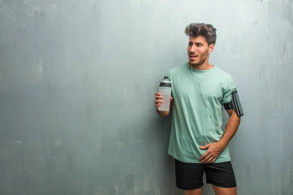 Young fitness man with sports bottle against a grunge wall very scared and afraid, wearing an armband with phone.