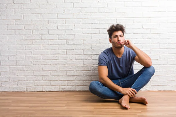 Young natural man sit on a wooden floor keeping a secret or asking for silence, serious face, obedience concept