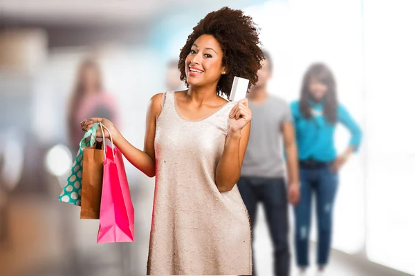 バック グラウンドでショッピング バッグやぼけの人のカードを保持している若い黒人女性 — ストック写真