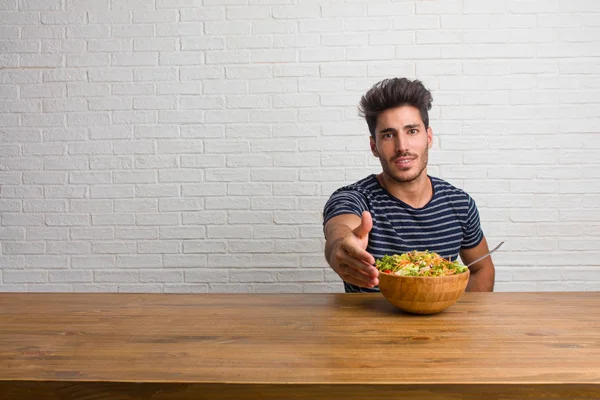 Young handsome and natural man sitting on a table reaching out to greet someone or gesturing to help, happy and excited. Eating a fresh salad.