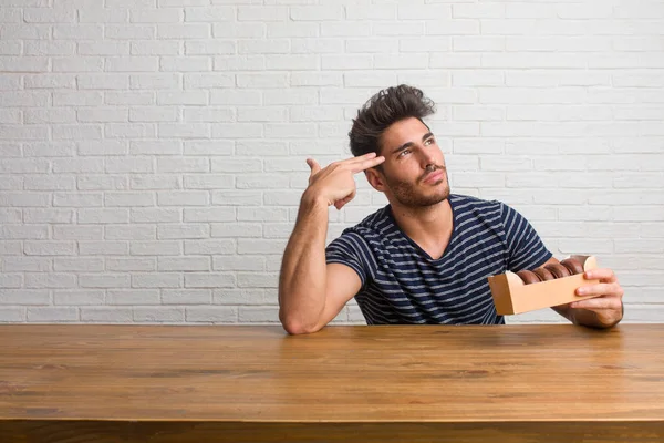 年轻英俊和自然的男人坐在桌子上做一个自杀的手势 感到悲伤和害怕形成一个手指的枪 吃巧克力甜甜圈 — 图库照片