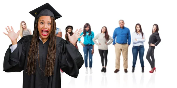 Young graduated black woman wearing braids screaming happy, surprised by an offer or a promotion, gaping, jumping and proud