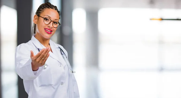 Portrait of a young black doctor woman inviting to come, confident and smiling making a gesture with hand, being positive and friendly