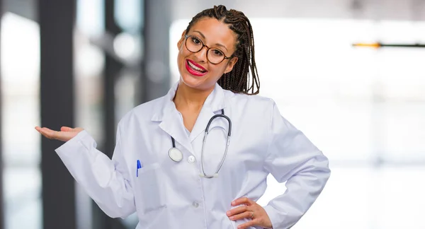 Portrait of a young black doctor woman holding something with hands, showing a product, smiling and cheerful, offering an imaginary object